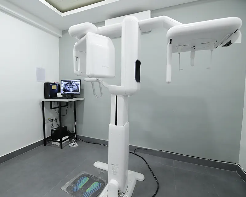 A dental x-ray machine in a dental office. The machine has a large white rectangular panel in the center for positioning patients. A black stand with a long arm extends out from the machine, holding a rectangular x-ray detector. There is a control panel with a computer screen on a stand next to the x-ray machine.